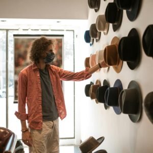 A man wearing a mask shops for hats in a boutique, touching various styles on display.