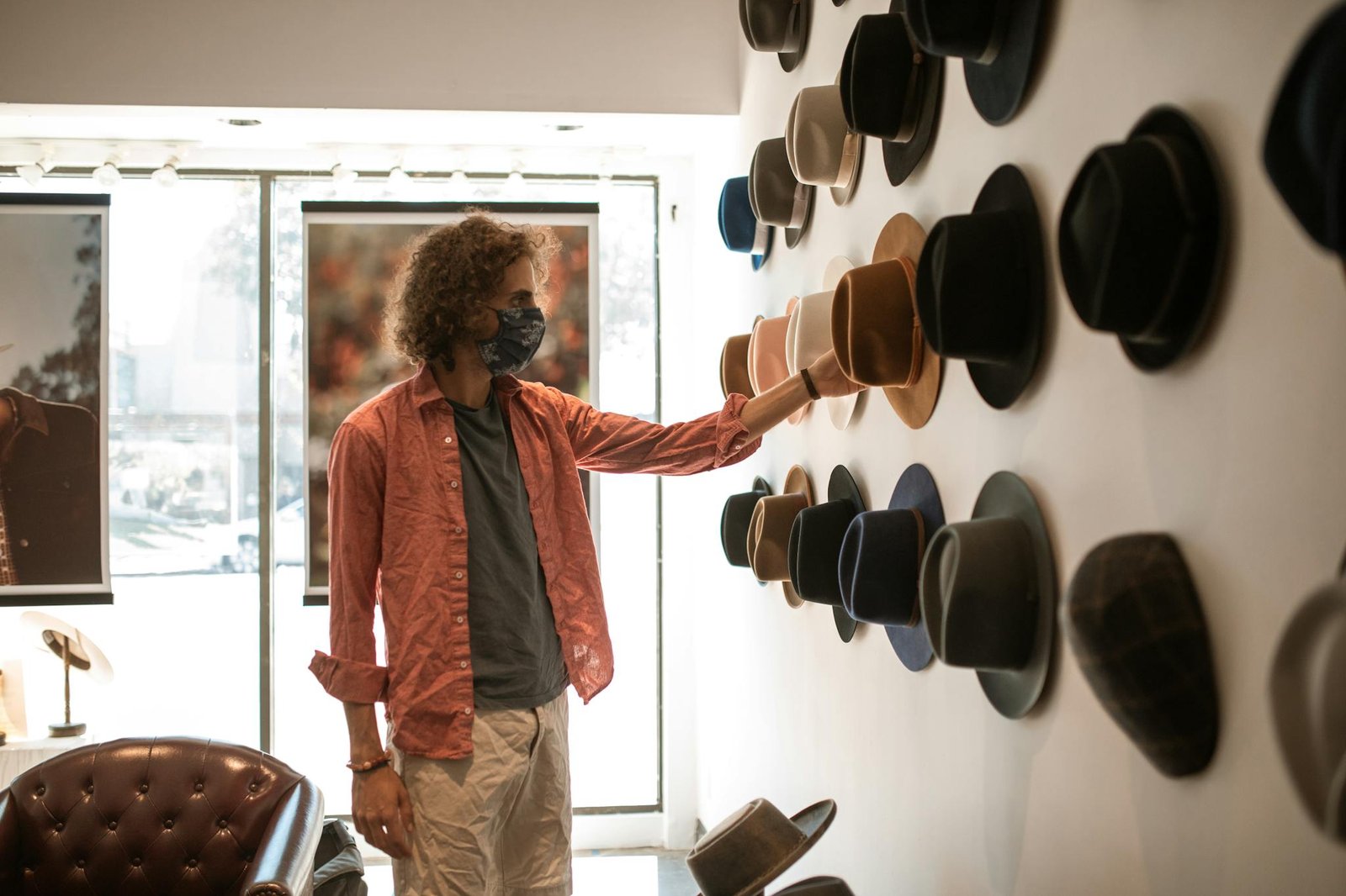 A man wearing a mask shops for hats in a boutique, touching various styles on display.